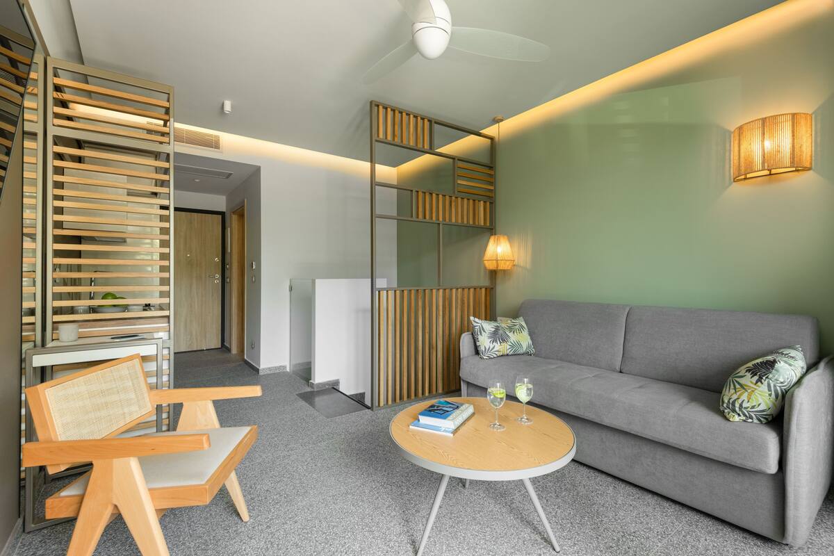 A modern living room with a grey sofa, a wooden chair, a round coffee table with a book and glasses, and a wall-mounted lamp.