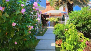 En sti, der fører til en bygning med et overdækket udendørs siddeområde, omgivet af frodig vegetation og blomstrende planter.