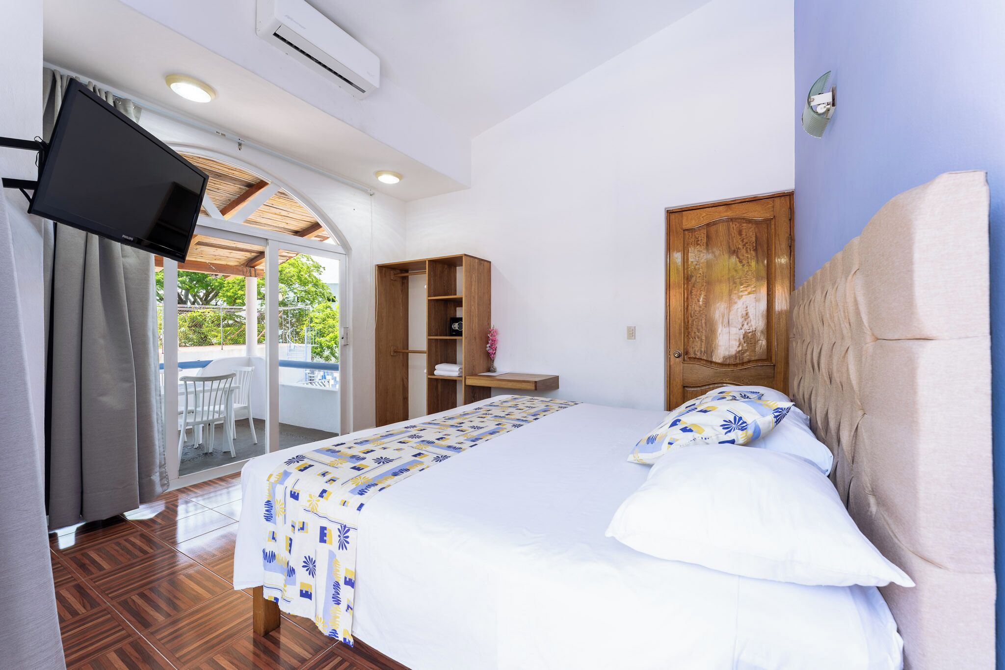 Un dormitorio con cama, televisor, armario de madera y puerta corrediza de vidrio que da a un espacio exterior.