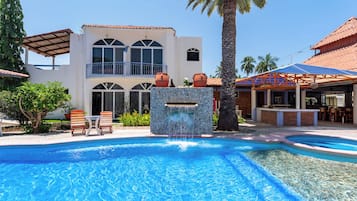 Una piscina amb cascada, un porxo cobert i un edifici de dues plantes amb grans finestres.