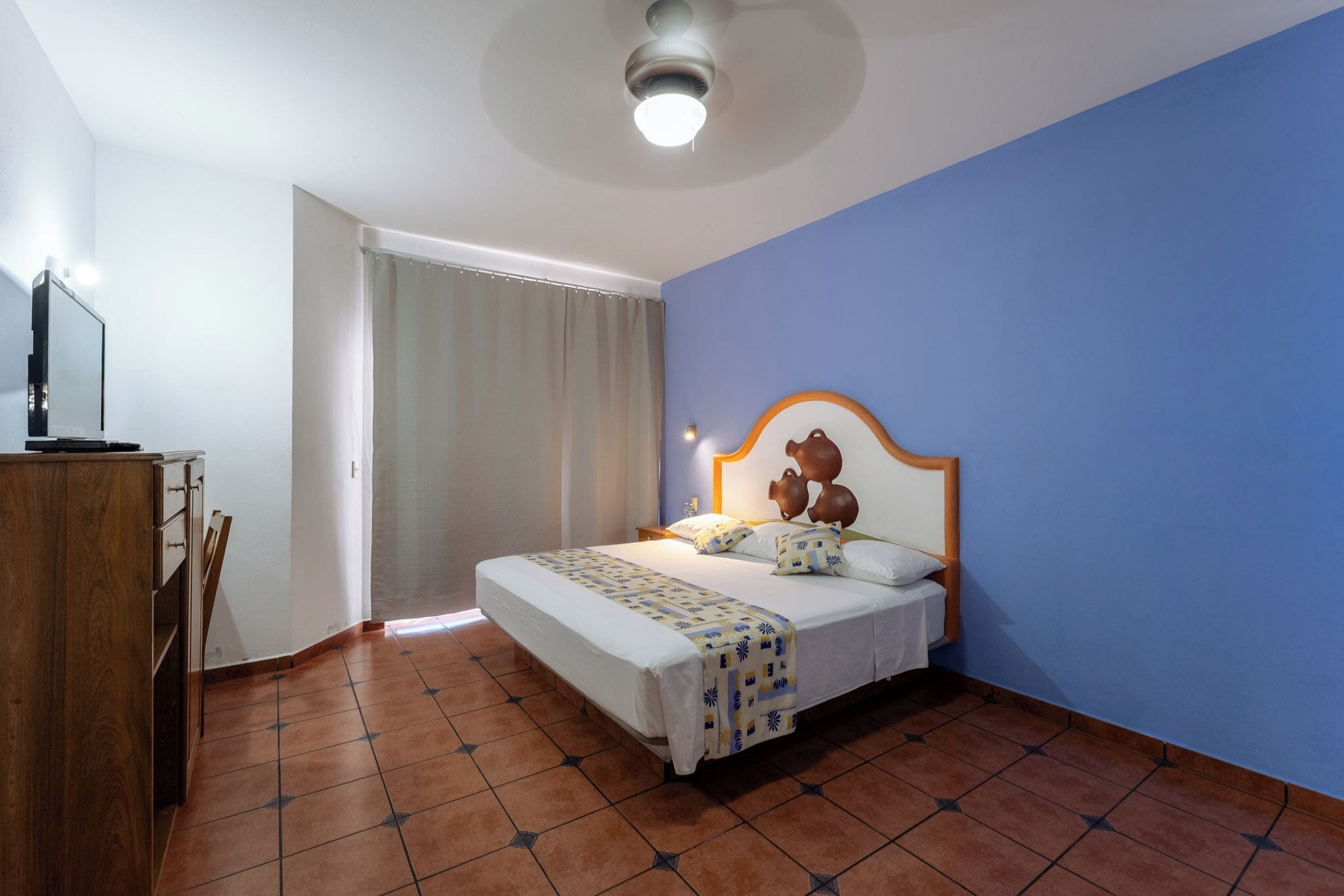 Un dormitorio con cama, cómoda de madera, ventilador de techo y ventana con cortinas.