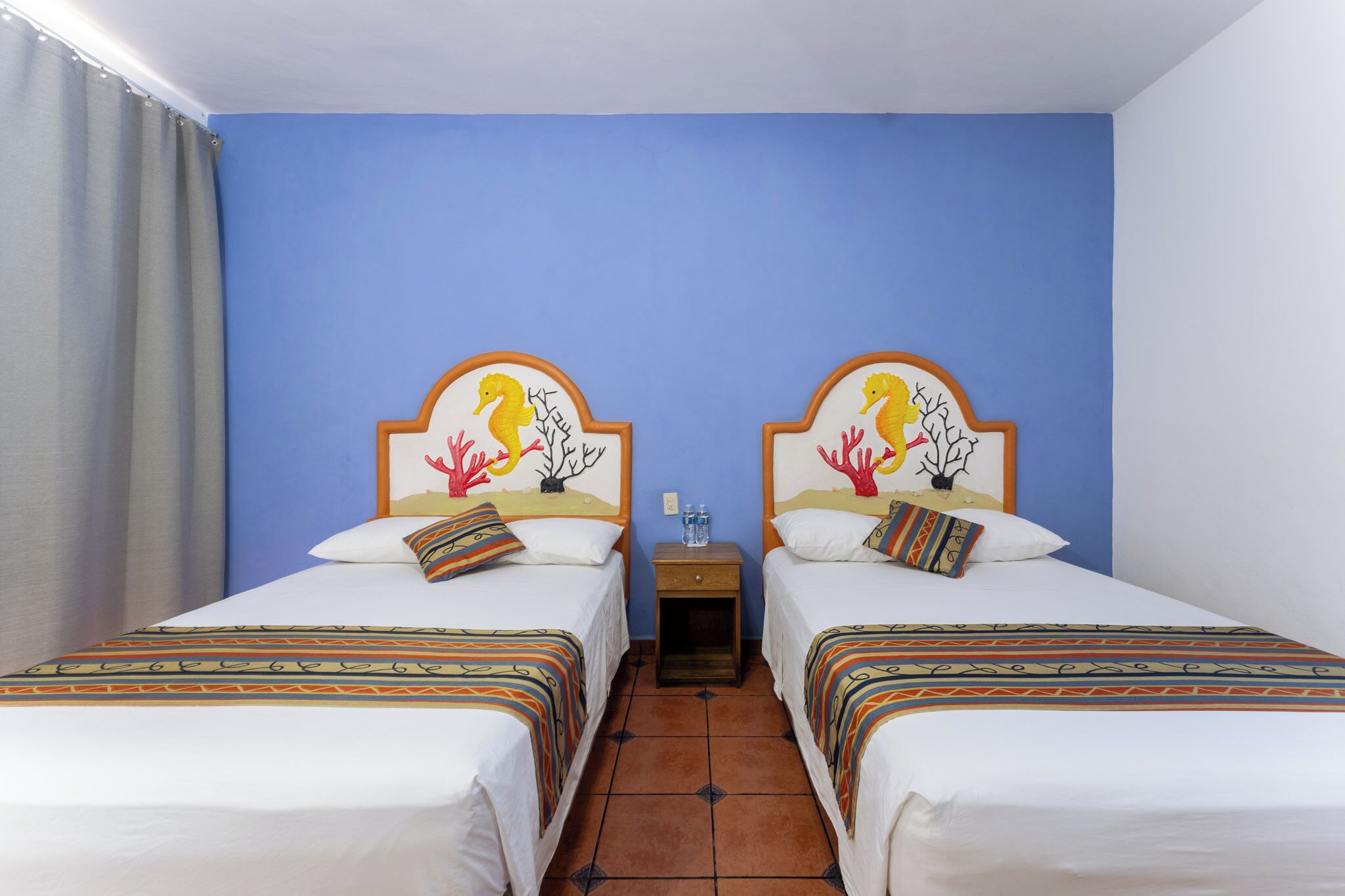 A hotel room with two beds, each with a unique headboard design, a small bedside table with a water bottle, and a curtain on the left side.