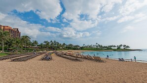 Ein Strand mit Liegestühlen, Sonnenschirmen, Palmen und einem Hotel im Hintergrund.