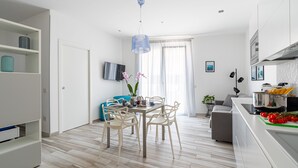 Una cocina moderna con armarios blancos, un comedor con una mesa y sillas, y una sala de estar con sofá y televisor.