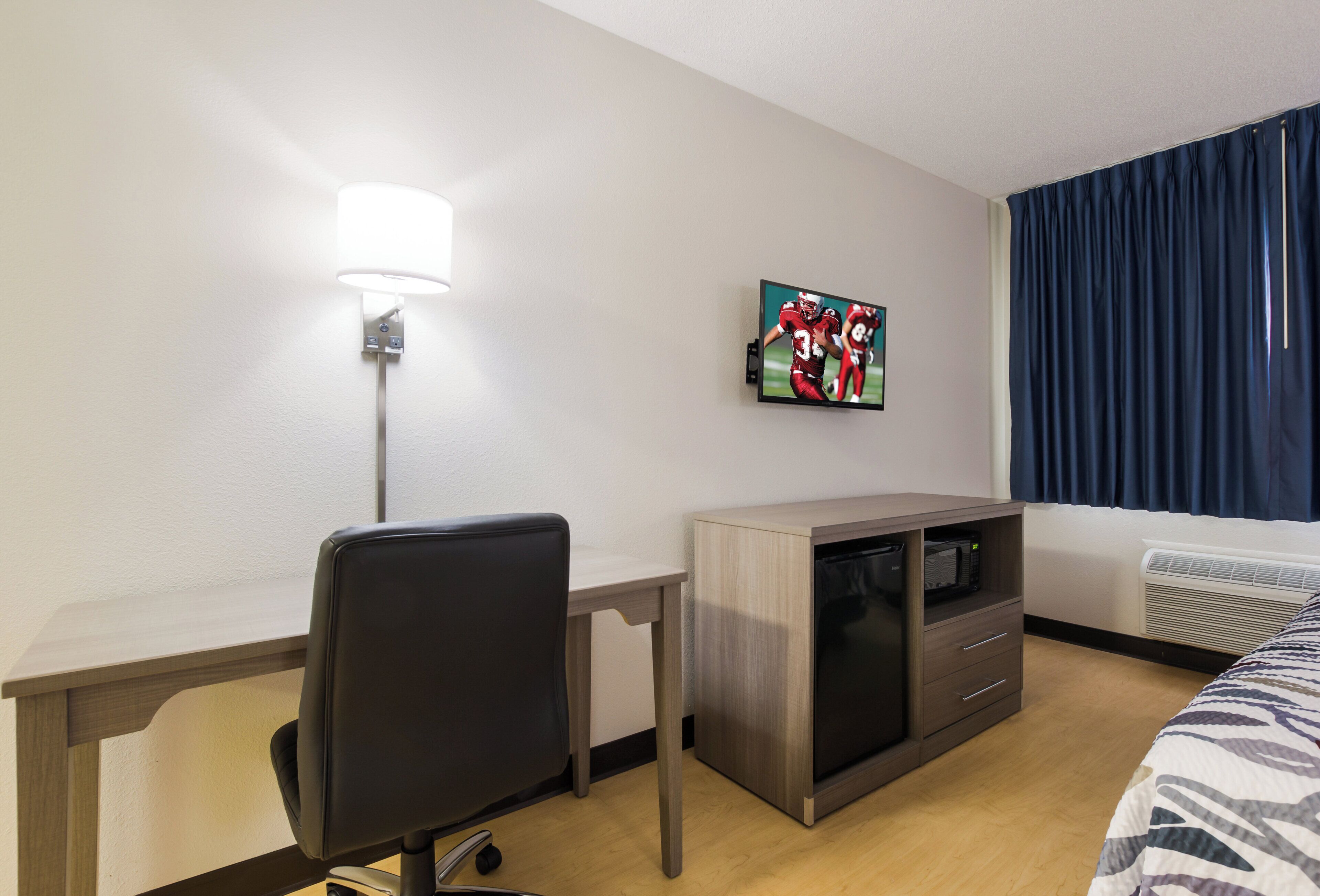 A hotel room with a desk, a chair, a television mounted on the wall, a microwave, and a bed with a patterned bedspread.