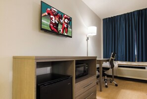 Habitación de hotel con una televisión de pantalla plana montada en la pared, un escritorio con una silla, un microondas y un calefactor.