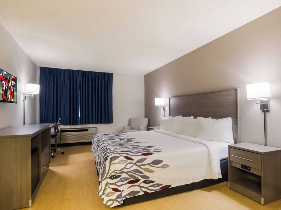 A hotel room with a bed, bedside tables, a desk, a chair, a TV, and a window with curtains.