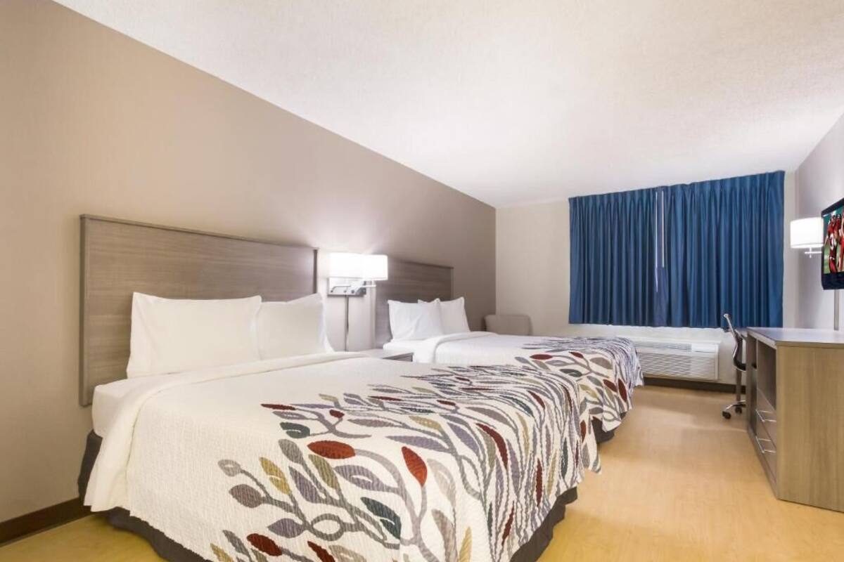 Quarto de hotel com duas camas, uma escrivaninha, uma cadeira, um abajur e uma janela grande com cortinas.