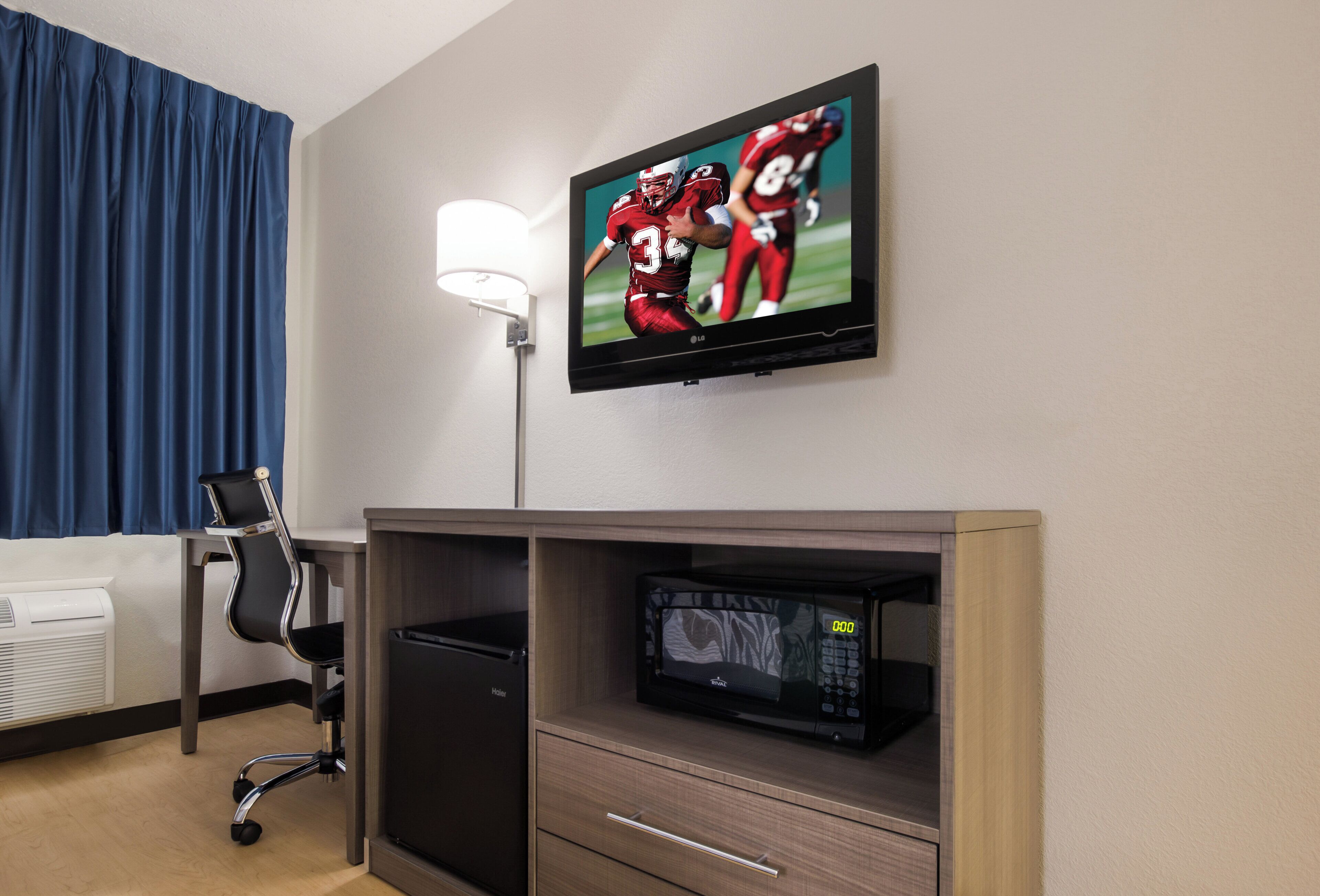 A hotel room with a flat-screen TV mounted on the wall, a desk with a chair, a microwave, and a heater.