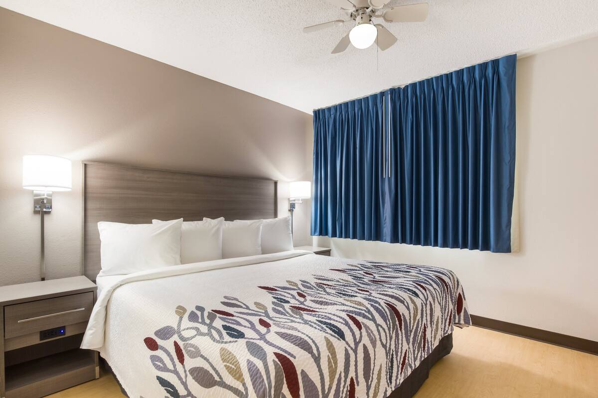 Quarto de hotel com cama, mesas de cabeceira, ventilador de teto e uma janela grande com cortinas azuis.