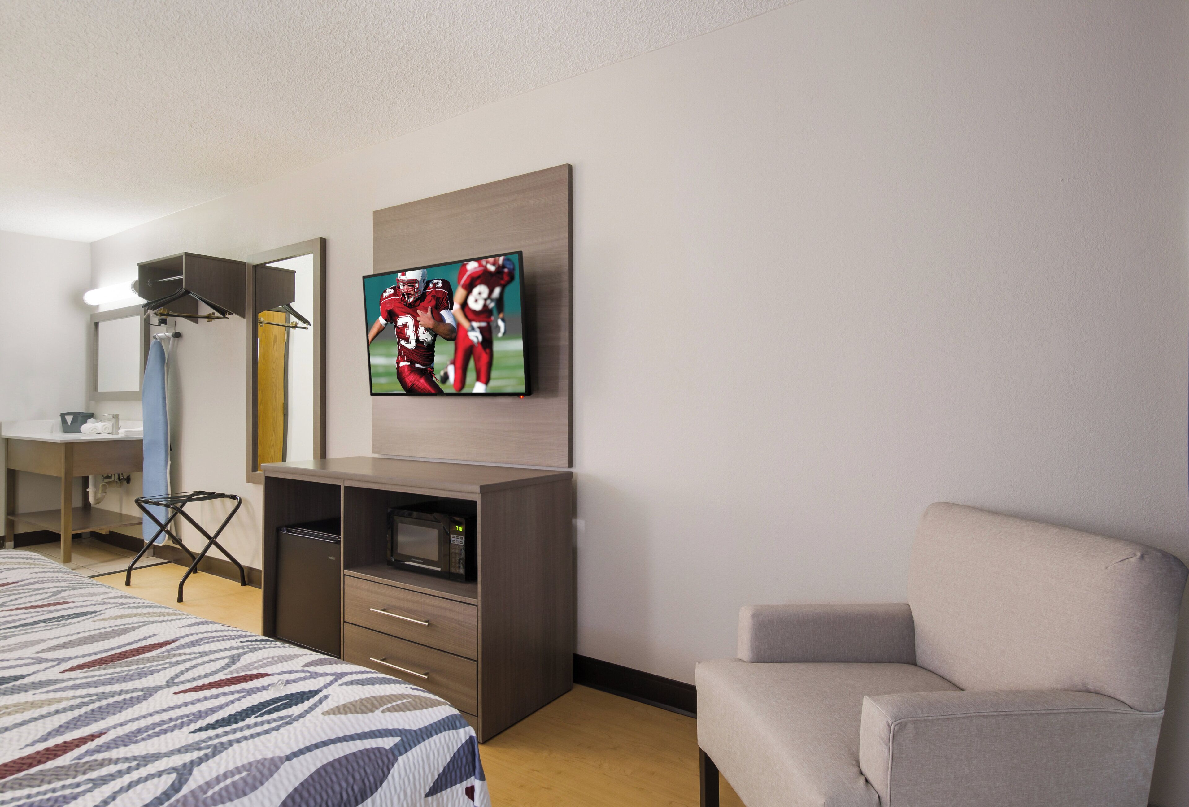A hotel room with a bed, a TV mounted on the wall, a desk with a microwave, and a chair.
