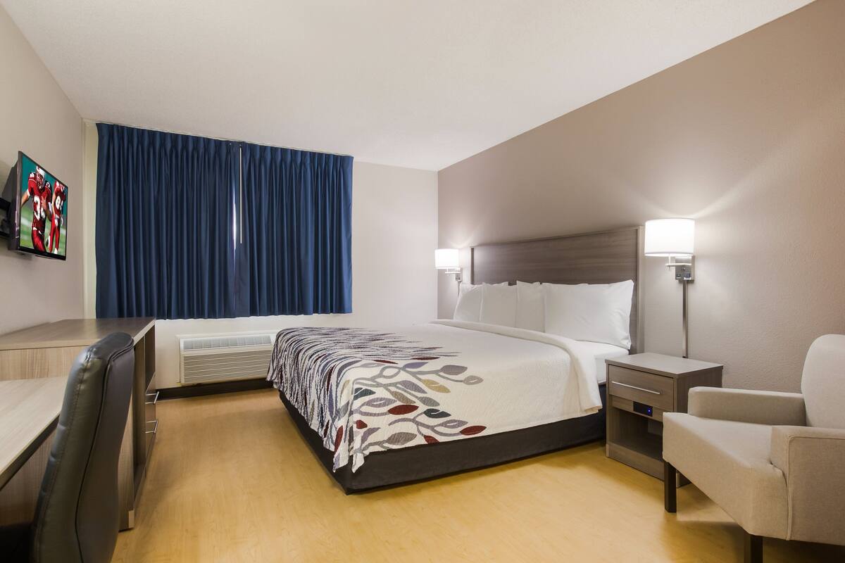 Quarto de hotel com cama, mesas de cabeceira, escrivaninha, cadeira e televisão.