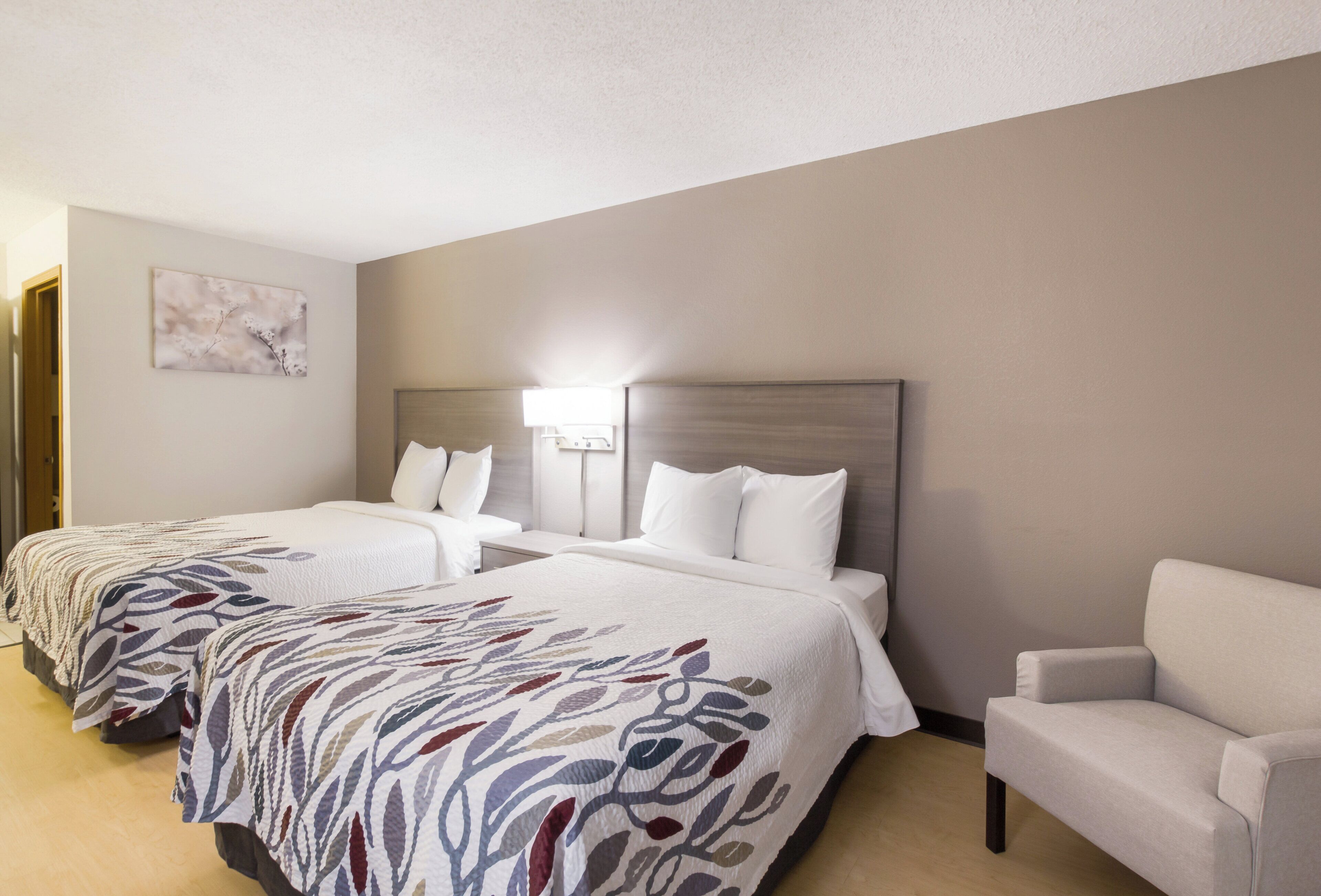A hotel room with two beds, a nightstand, a chair, and a wall-mounted lamp.