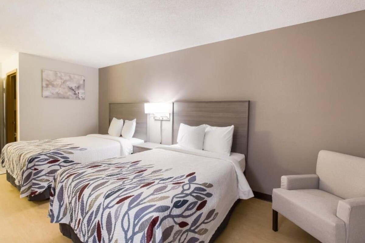 A hotel room with two beds, a nightstand, a lamp, and a chair.