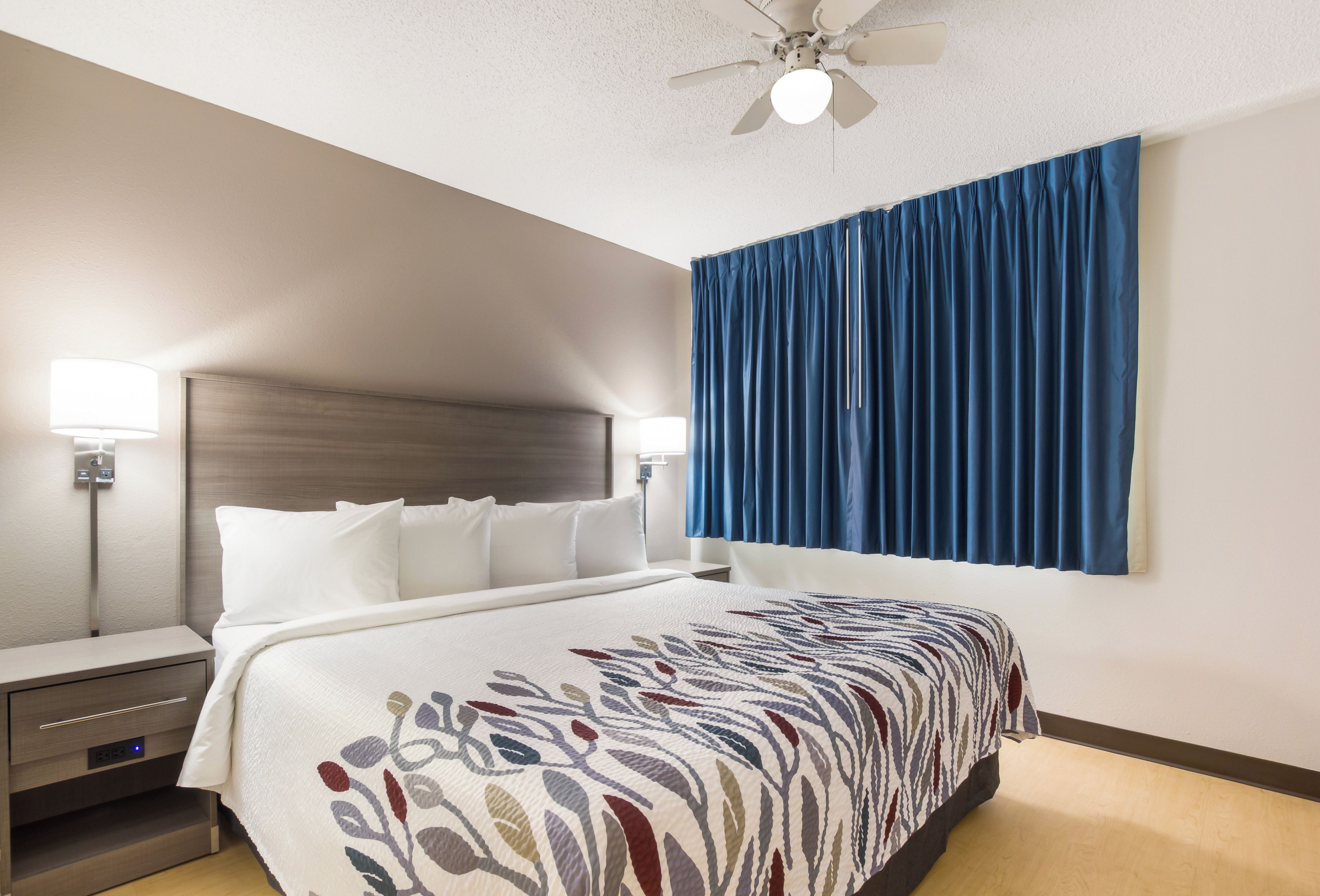 A hotel room with a bed, bedside lamps, a ceiling fan, and a large window with blue curtains.