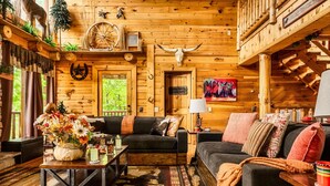 A rustic cabin interior with wooden walls, a black leather sofa, a coffee table with a floral arrangement, and a cowhide rug.