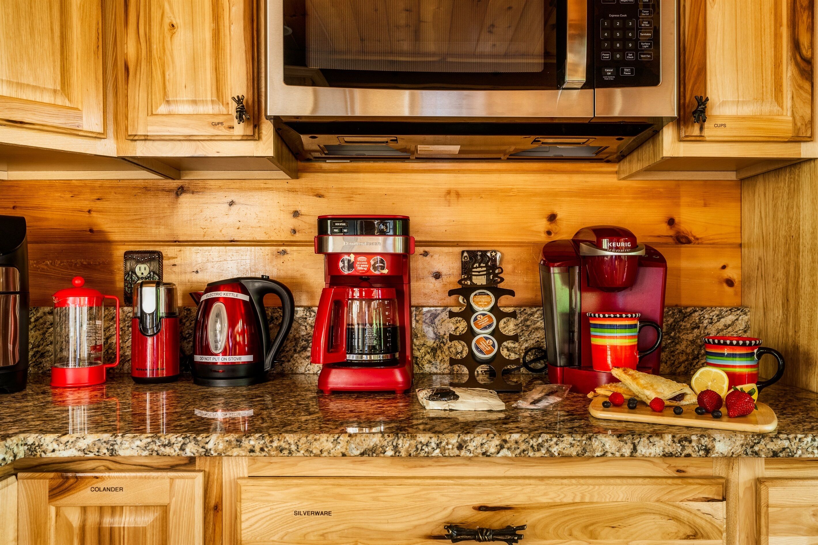 A kitchen with wooden cabinets, a microwave, a coffee maker, a kettle, and a fruit platter.
