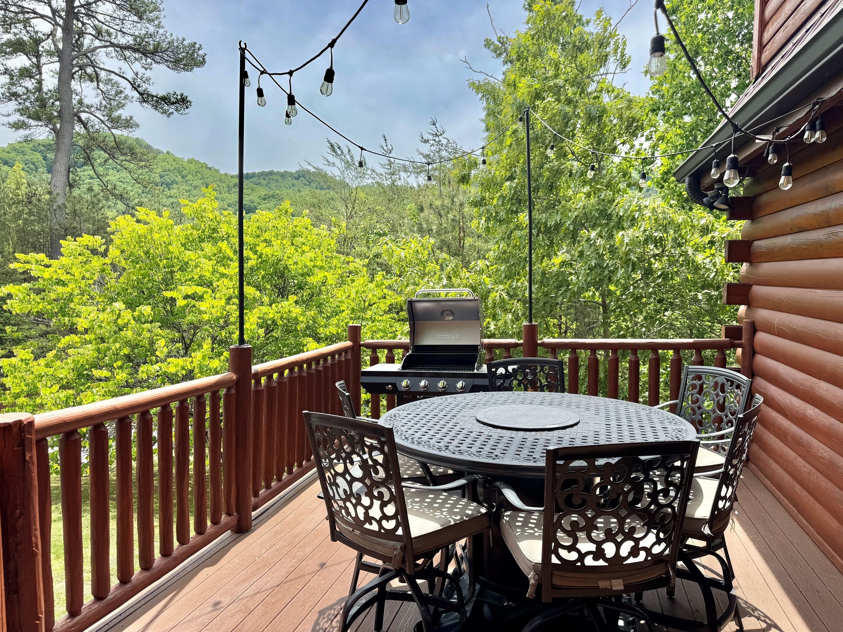 A wooden deck with a round table and chairs, a grill, and string lights.