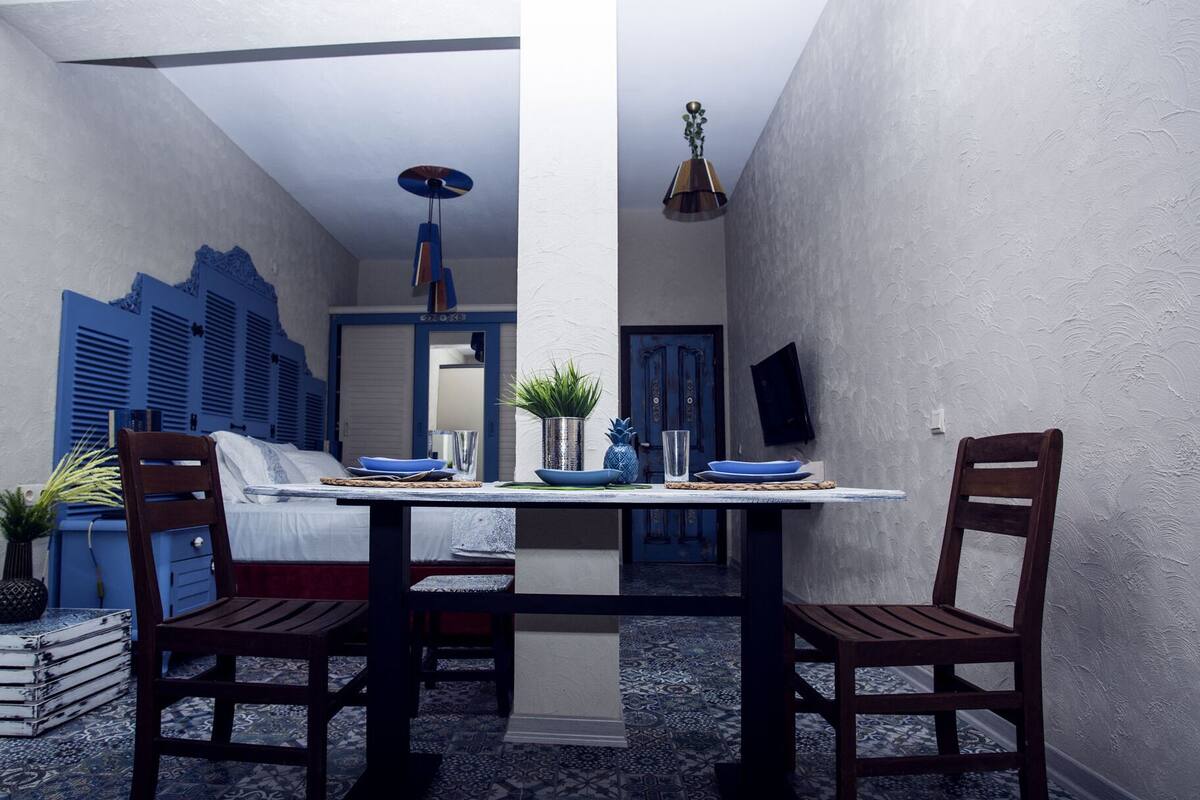 A dining area with a table set for two, chairs, a potted plant, and a blue door in the background.