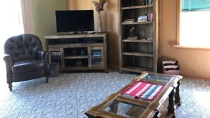 A living room with a leather armchair, a flat-screen TV on a wooden stand, a bookshelf, and a glass coffee table.