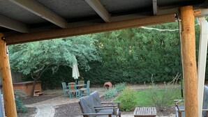 Een overdekt terras met bakstenen vloer, houten meubilair en uitzicht op een weelderige tuin.