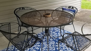 Una mesa metálica redonda con cuatro sillas, una cesta tejida y una alfombra azul estampada sobre un patio de concreto.