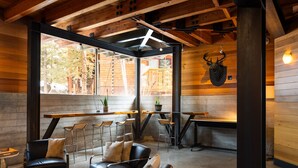 Um interior rústico de madeira com área de estar, incluindo sofás de couro e cadeiras de metal, espaço de bar com banquetas e uma grande janela com vista para o exterior.