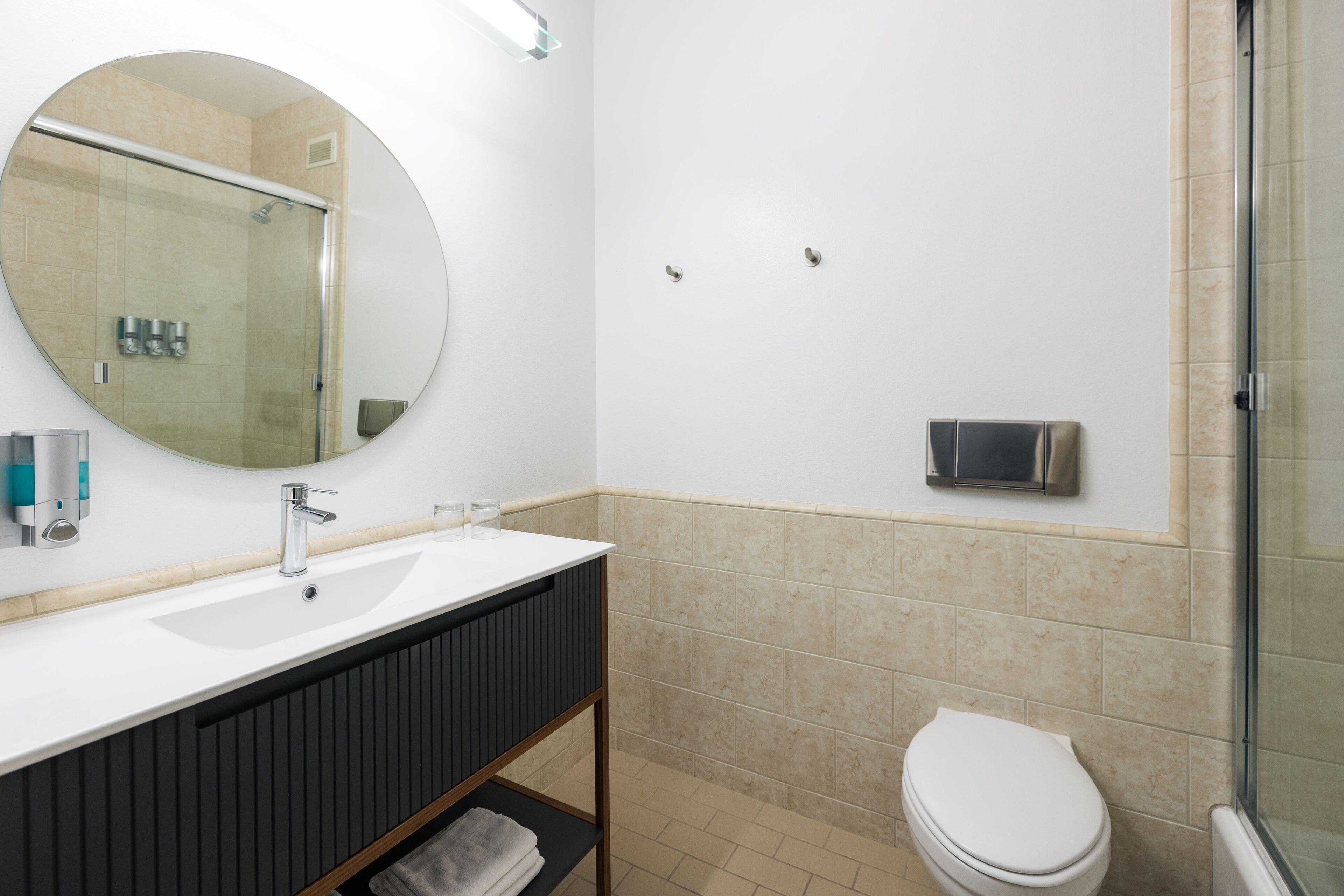 A bathroom with a white sink, dark vanity, a round mirror, a toilet, and a glass shower enclosure.