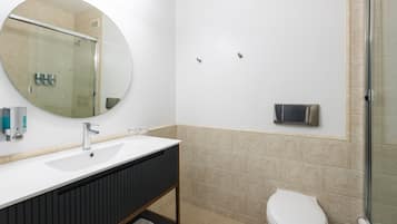 A bathroom with a white sink, dark vanity, a round mirror, a toilet, and a glass shower enclosure.