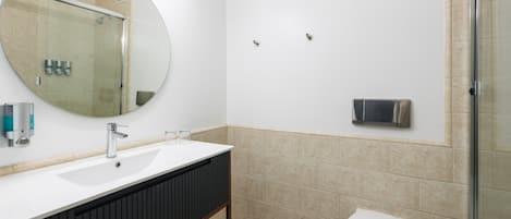 Un baño con lavamanos blanco, tocador oscuro, espejo redondo, inodoro y cabina de ducha con mampara de vidrio.