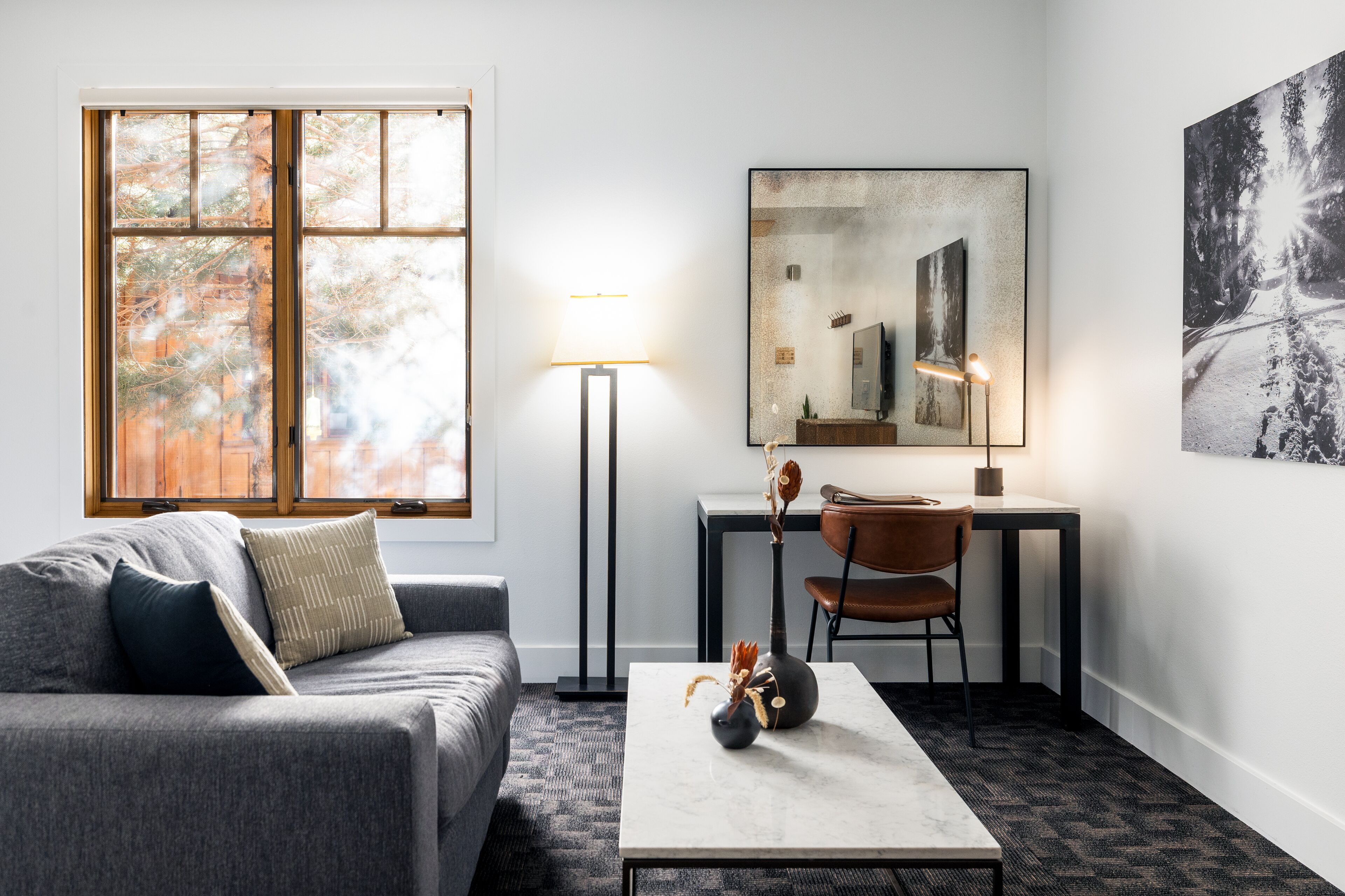 Una sala de estar moderna con un sofá gris, una mesa de centro con adornos, un escritorio con una silla, una lámpara de pie y un cuadro grande en la pared.