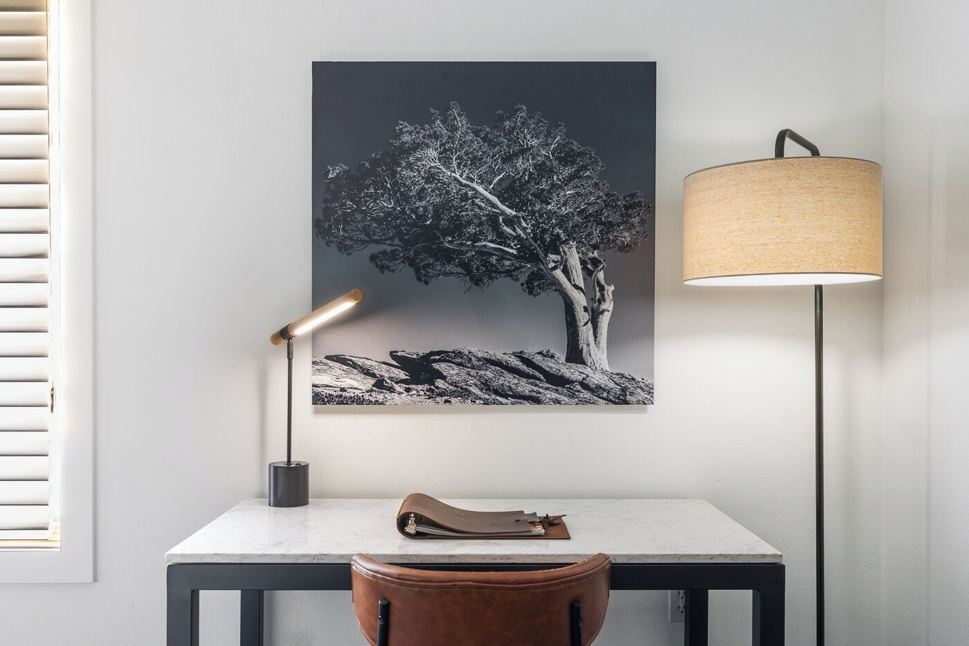 A desk with a lamp, a chair, a leather bag, and a framed black and white tree photograph on the wall.