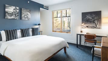 Quarto de hotel com uma cama grande, uma escrivaninha e uma cadeira. Há duas janelas e quadros nas paredes.