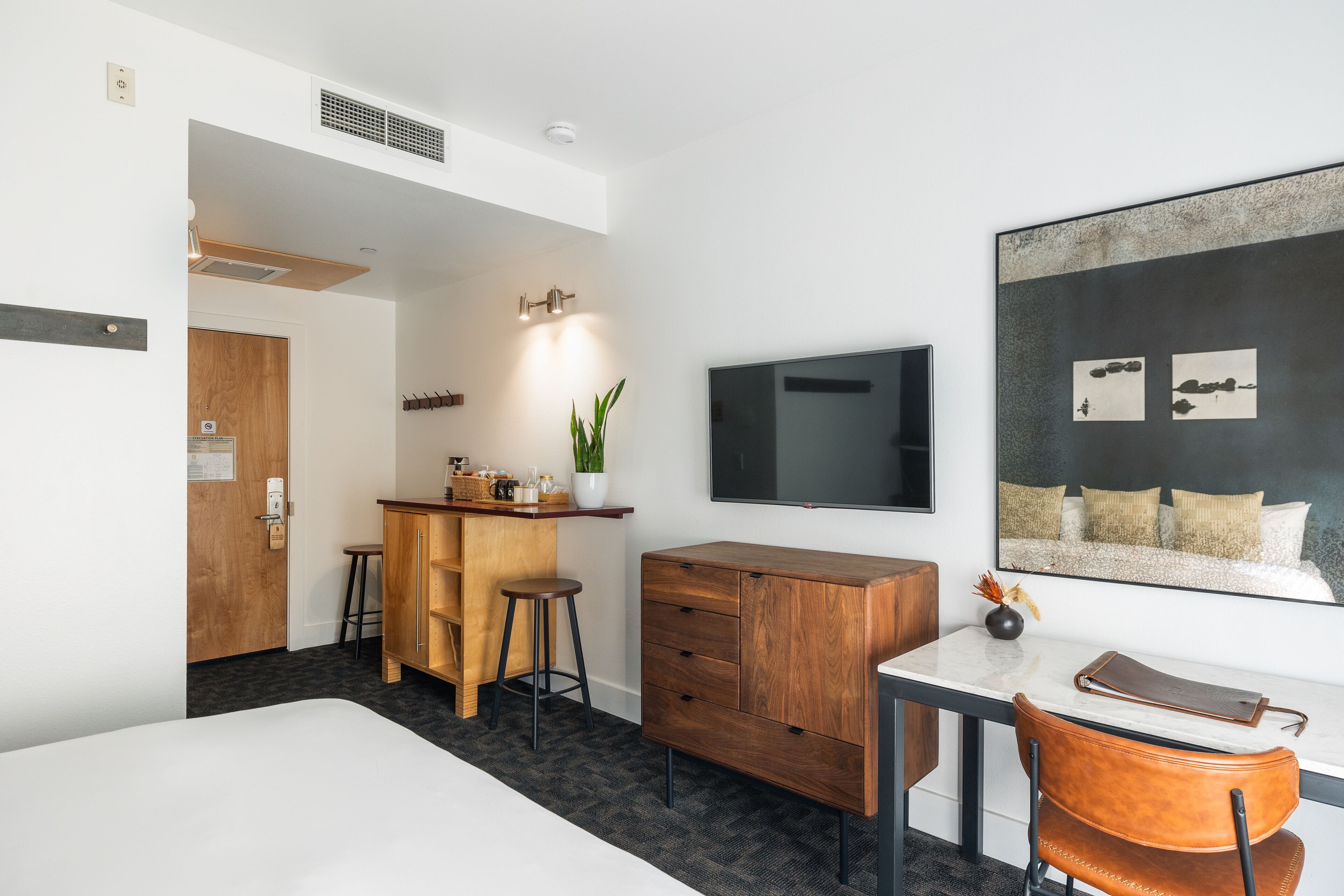 Una habitación de hotel moderna con una cama, un escritorio con una silla, un aparador de madera, una zona de bar con taburetes, una planta en maceta y una obra de arte grande enmarcada en la pared.