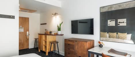 Una habitación de hotel moderna con una cama, un escritorio con una silla, un aparador de madera, una zona de bar con taburetes, una planta en maceta y una obra de arte grande enmarcada en la pared.