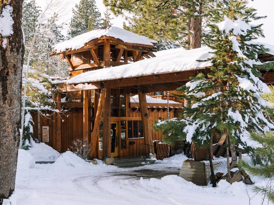 Une cabane en bois dont le toit est recouvert de neige, entourée d’arbres et bordée d’un sentier enneigé.