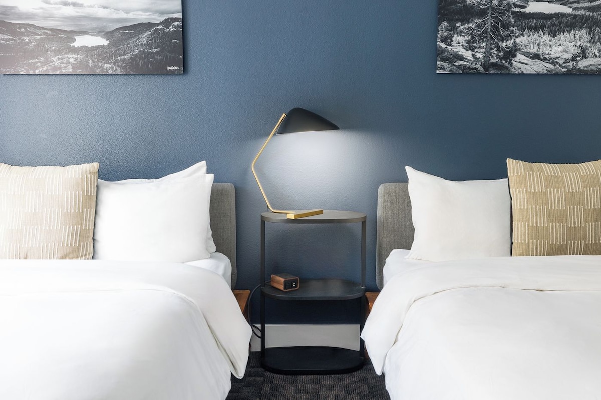 A hotel room with two beds, a bedside table with a lamp, and two framed pictures on the wall.