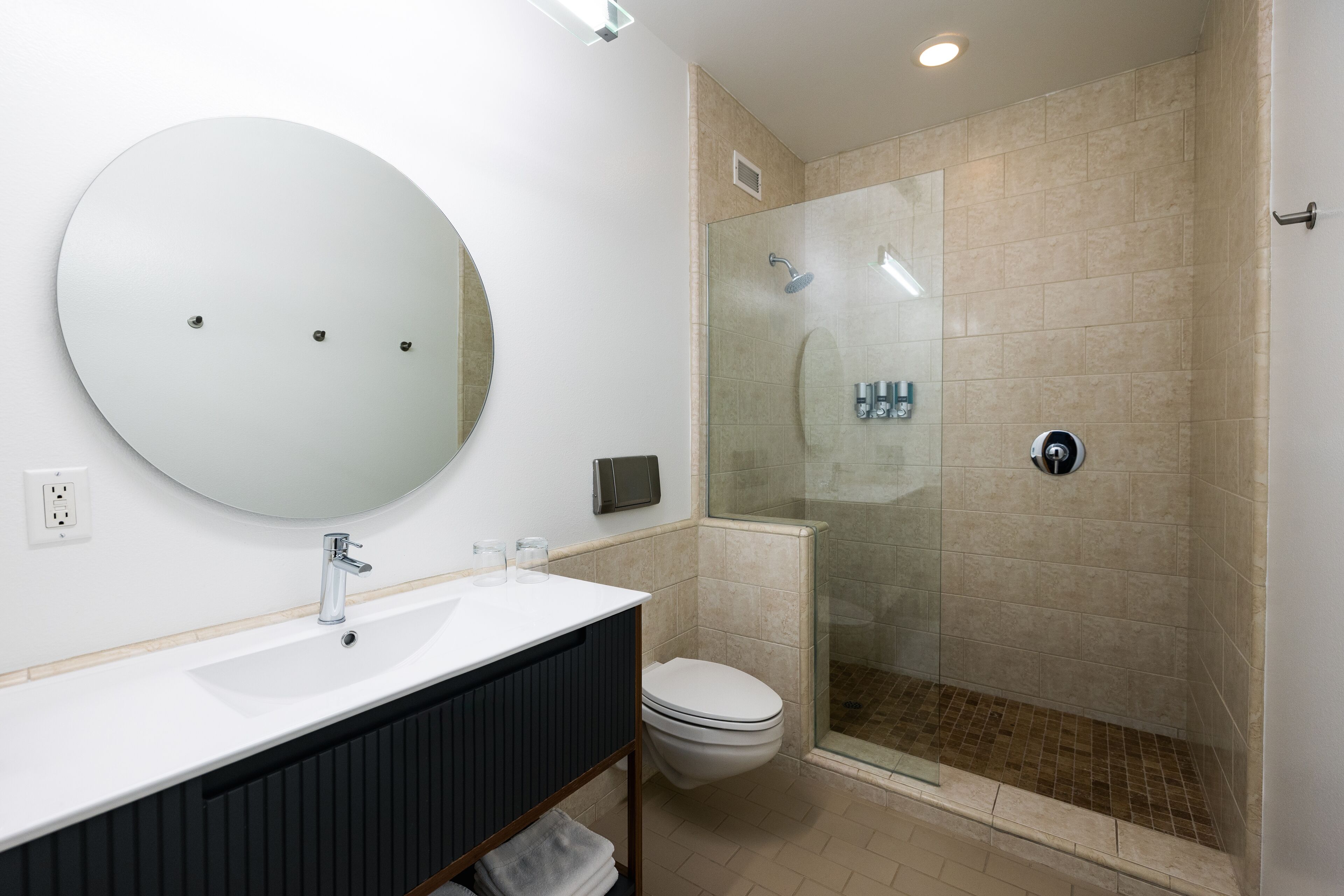 Un baño moderno con un espejo redondo, un lavabo blanco, un inodoro y una zona de ducha con mampara de cristal.