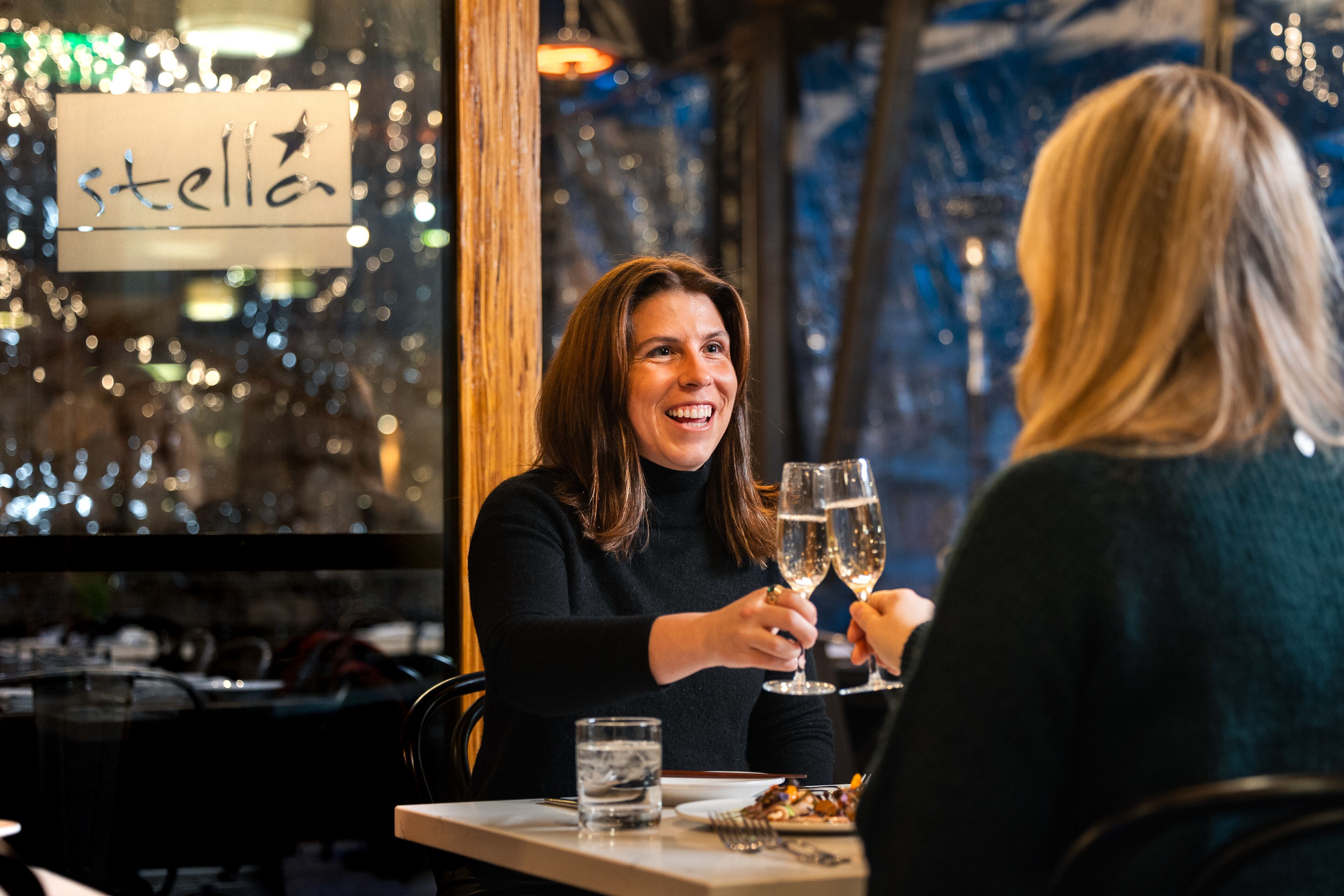 A restaurant interior with a sign reading "stella" on the window, a table with two people toasting, and a glass of water on the table.