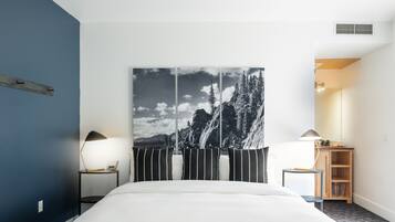 Ein Hotelzimmer mit einem großen Bett, einem Nachttisch mit Lampe, einem Holzschrank und einem Wandbild einer Berglandschaft.