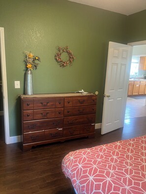 A room with a wooden dresser, a door, a patterned bedspread, and a wall with a wreath.
