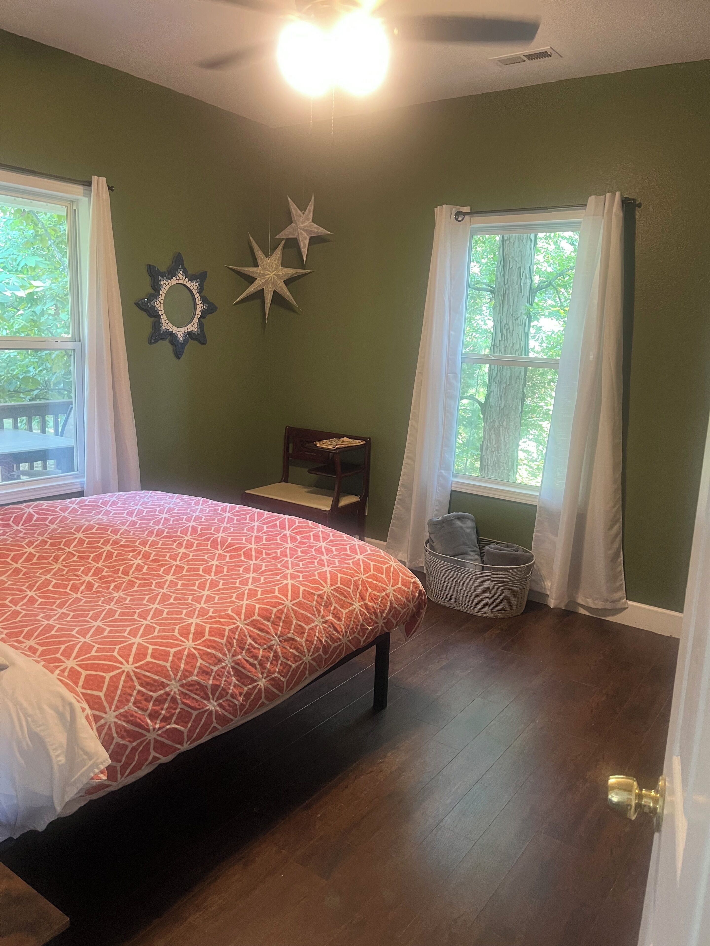 Une chambre à coucher avec un grand lit, deux fenêtres avec des rideaux, une table de chevet, une chaise et des éléments décoratifs accrochés au mur.