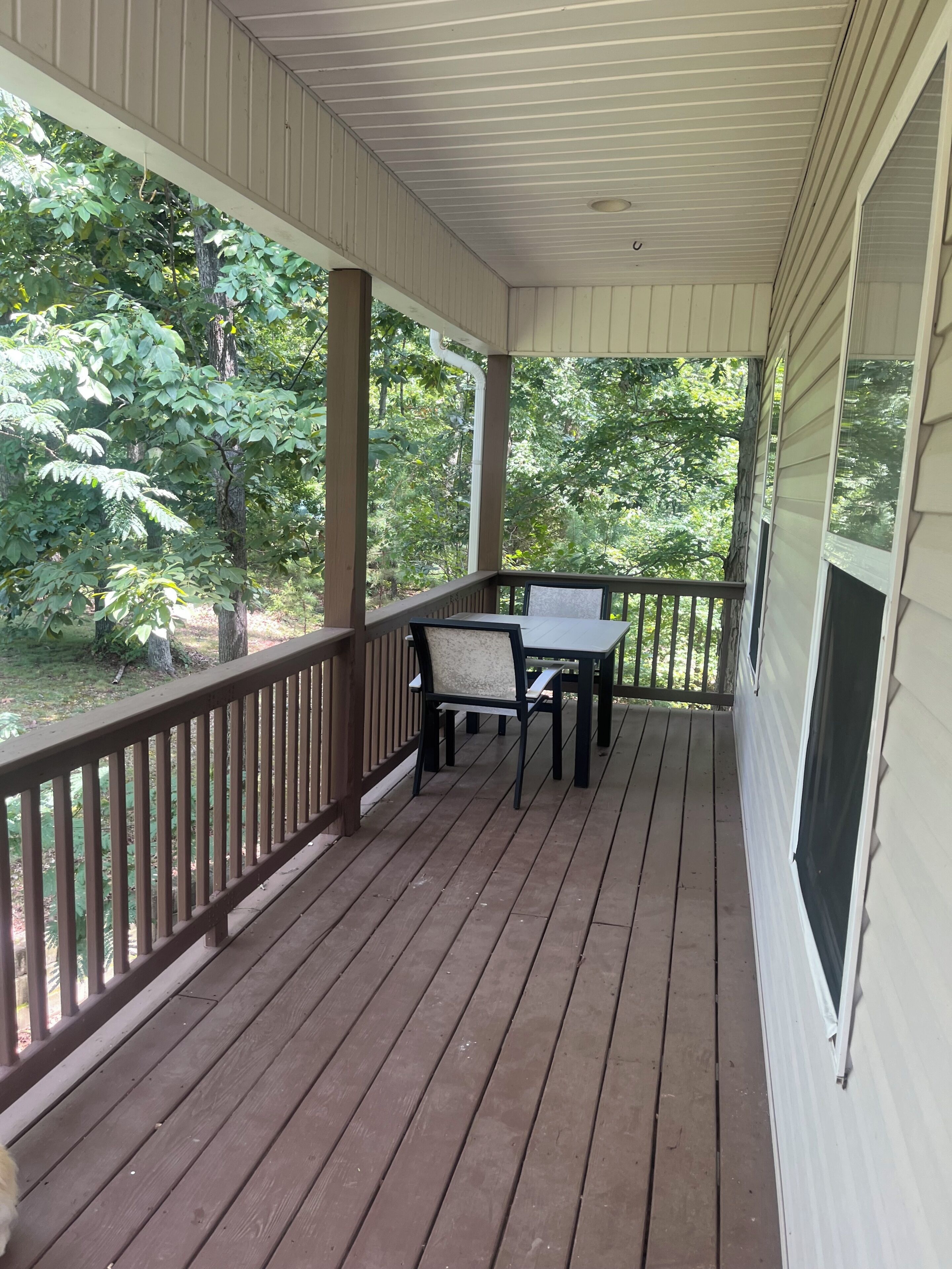 A covered porch with a table and chairs, surrounded by wooden railings and a view of greenery.