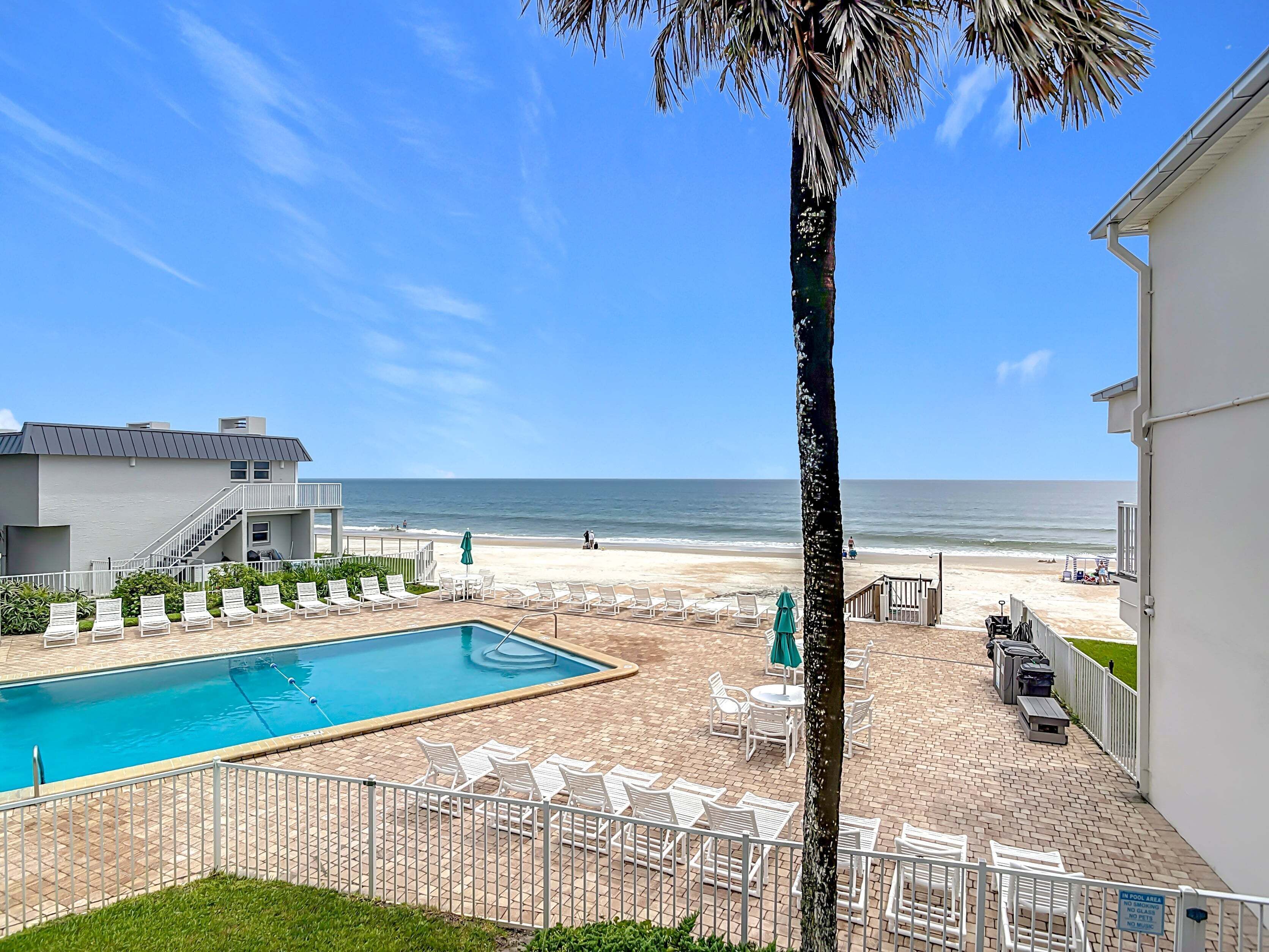 A pool area with lounge chairs, a palm tree, and a view of the beach and ocean.