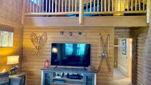 A log cabin interior with a TV on a wooden stand, a balcony, and ski equipment.
