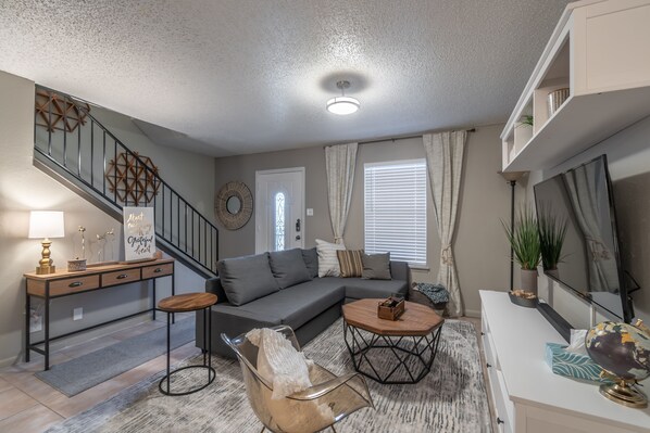 A living room with a gray sofa, a wooden coffee table, a glass chair, a console table with a lamp, and a staircase with a geometric wall decoration.