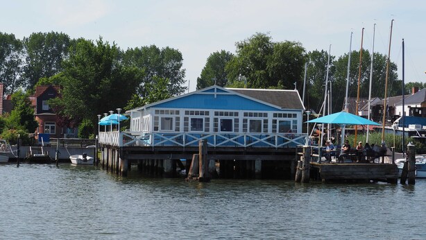 A docked boat with a blue building on stilts, a pier, and a row of houses in the background. - Studio Apartment Arnis - Apartments Landhaus Siemens in Kappeln (Kappeln)