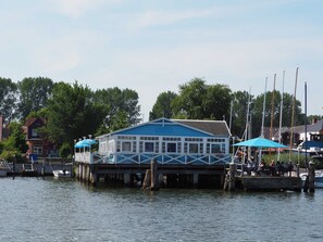 Un barco amarrado junto a un edificio azul sobre pilotes, un muelle y una fila de casas al fondo.