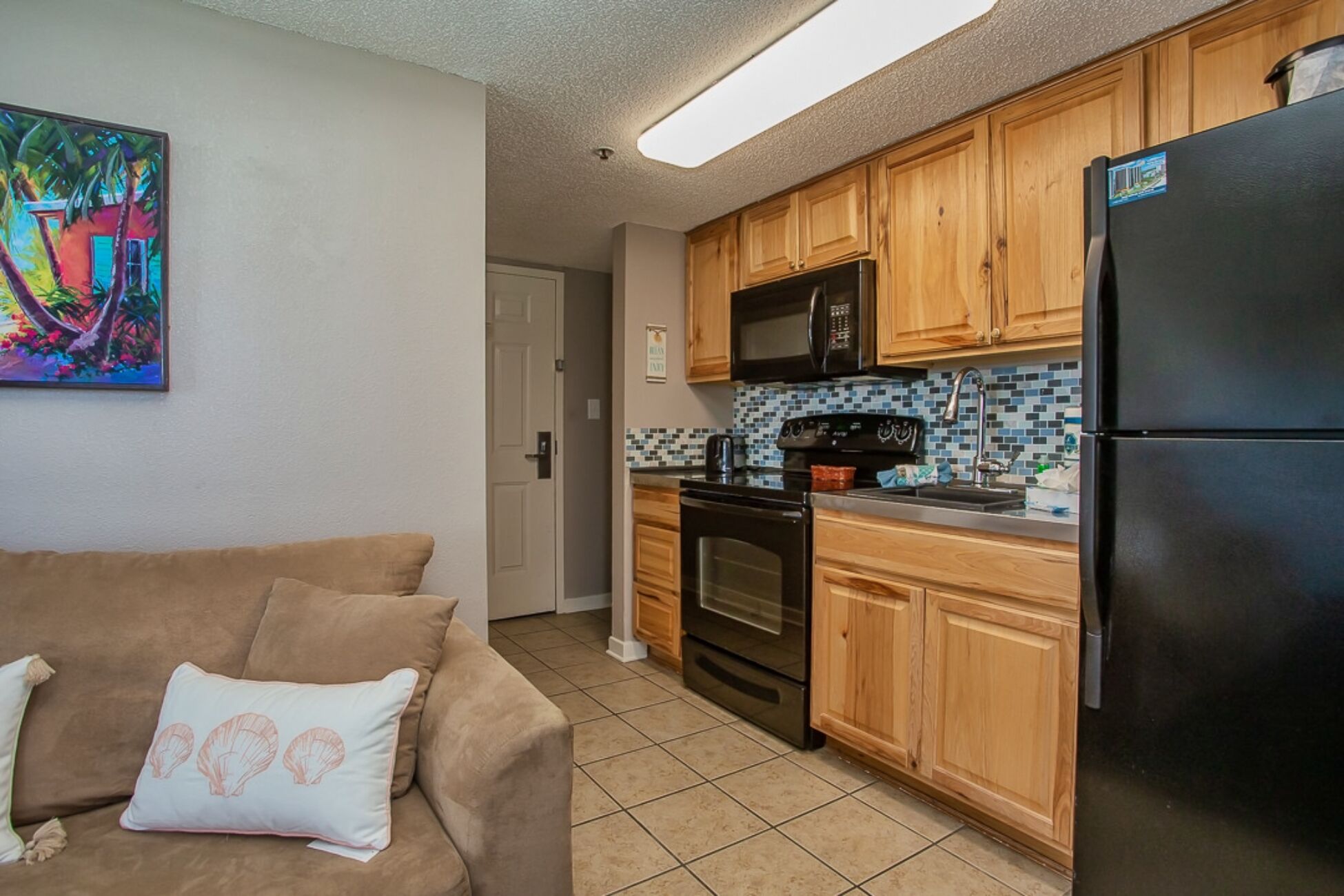 A compact kitchen with wooden cabinets, a black refrigerator, a microwave, and a tiled backsplash.