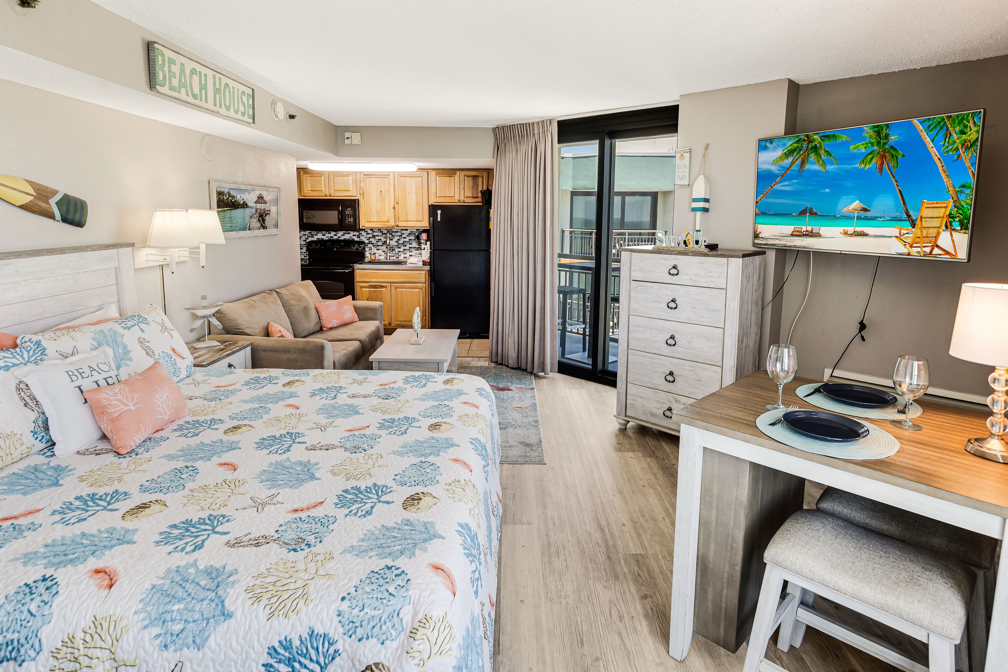 Ein Hotelzimmer mit Bett, Essbereich, Küche und Balkon. Ein Fernseher zeigt eine Strandansicht.